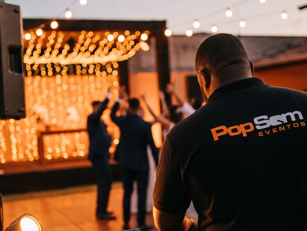Técnico da Pop Som de costas em uma festa, com cordões de luzes ao fundo
