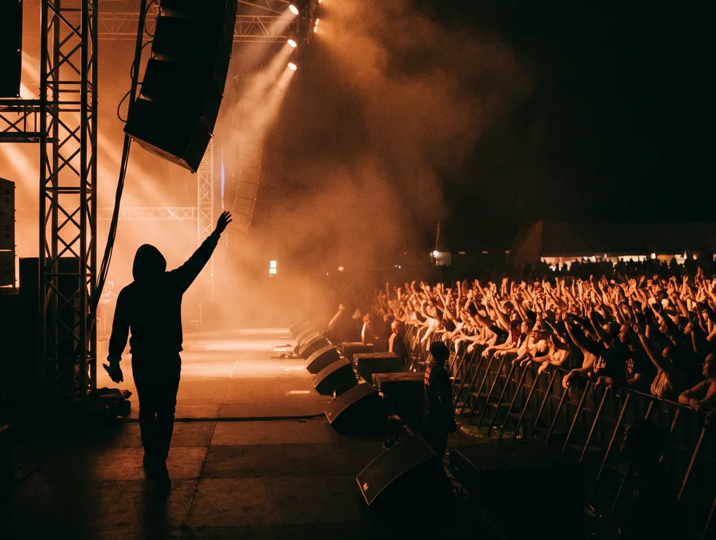 Artista de costas no palco, diante do público, entre fumaça e luzes alaranjadas